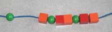 Une suite de perles forme un collier. 3 éléments sont répétés 3 fois, sphère verte, cube rouge, cube orange. Au début du collier un cube rouge est manquant et il y a un espace vide.