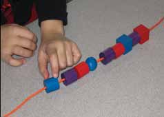 Les mains d' un enfant désignent la première forme d'une suite à motifs répétés : cylindre bleu, cylindre violet, cylindre rouge, répété 3 fois.