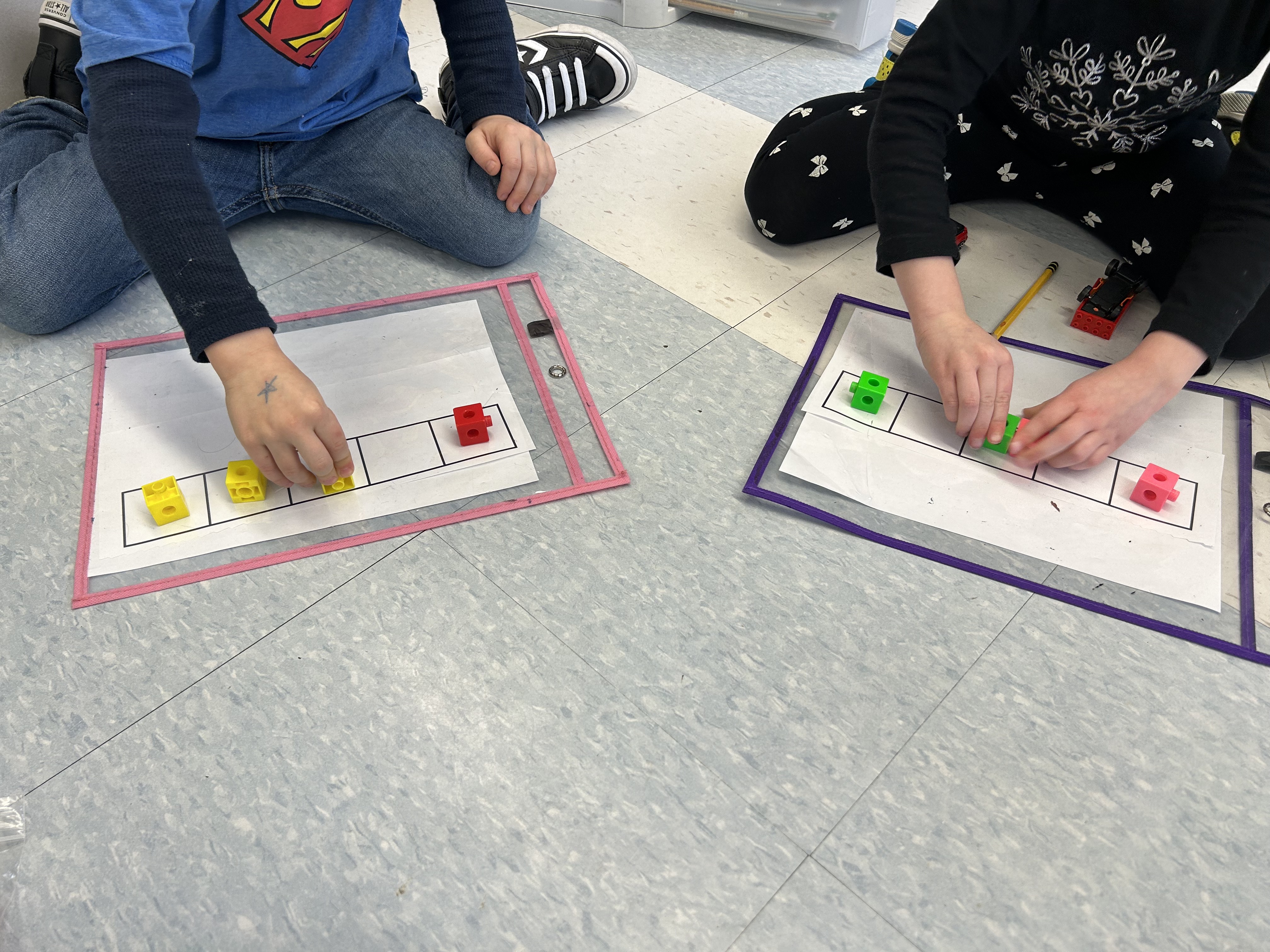 En salle de classe, deux jeunes garçons assis en tailleur jouent à empiler des jetons sur des cartes à cases.
