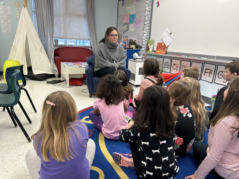 A classroom with young students. In front sits a teacher with her students sitting on a mat in front of her.