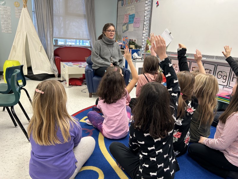 A classroom with young students. In front sits a teacher with her students sitting on a mat in front of her. Several students have their hands up.