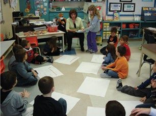 Les élèves sont assis, en cercle, par terre, ils ont de grandes feuilles devant eux. L’enseignante est avec eux. Elle regarde le travail qu’une élève présente.